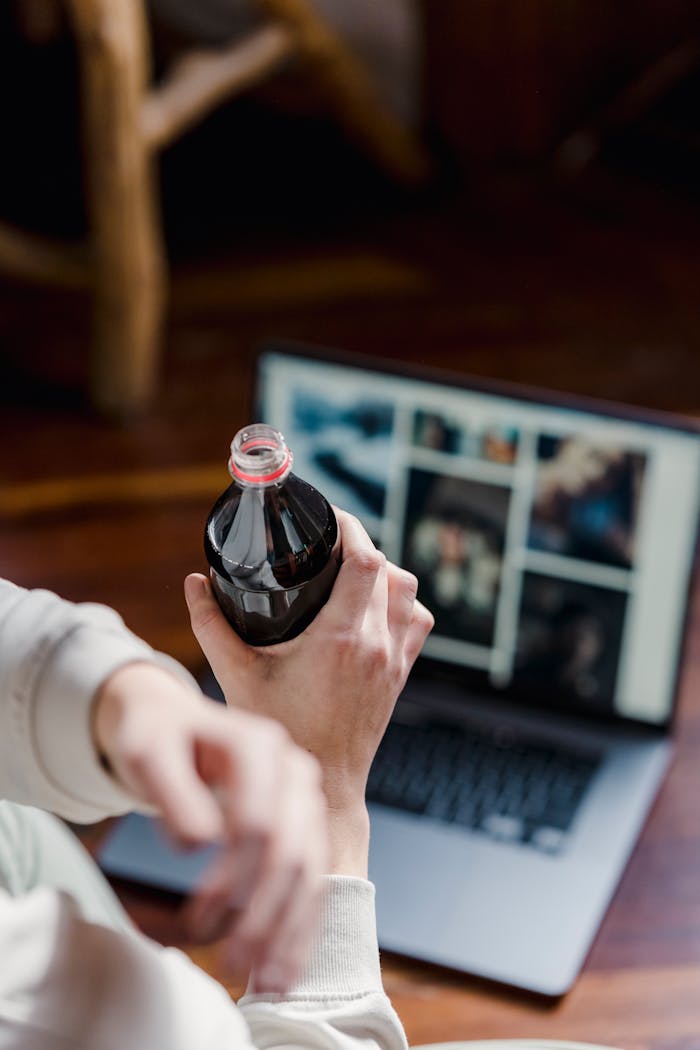Unrecognizable person with opened bottle of refreshing soda in hand sitting on floor with laptop on blurred background at home